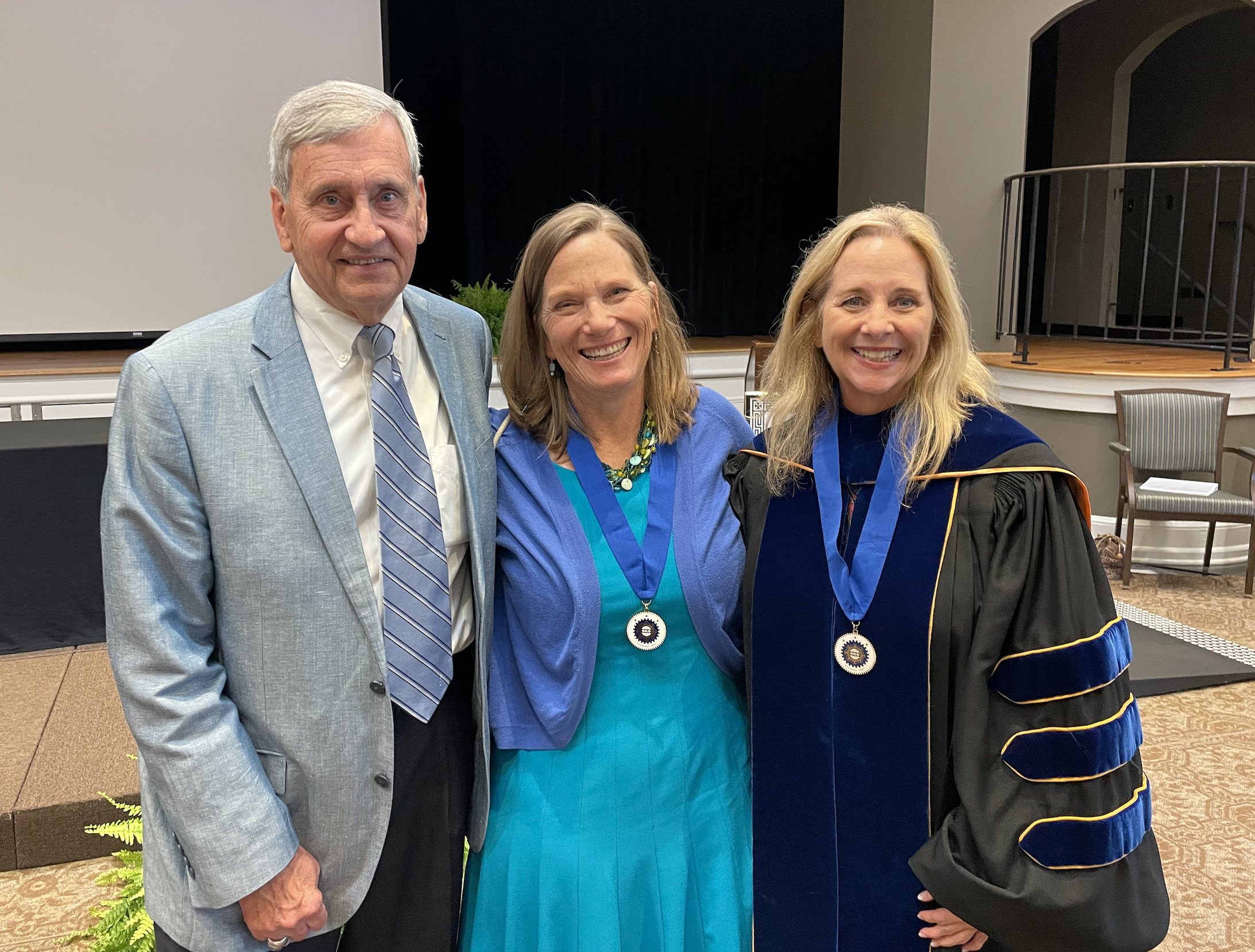 Three people smile and pose together at an academic event, with two wearing medallions and one in graduation regalia.